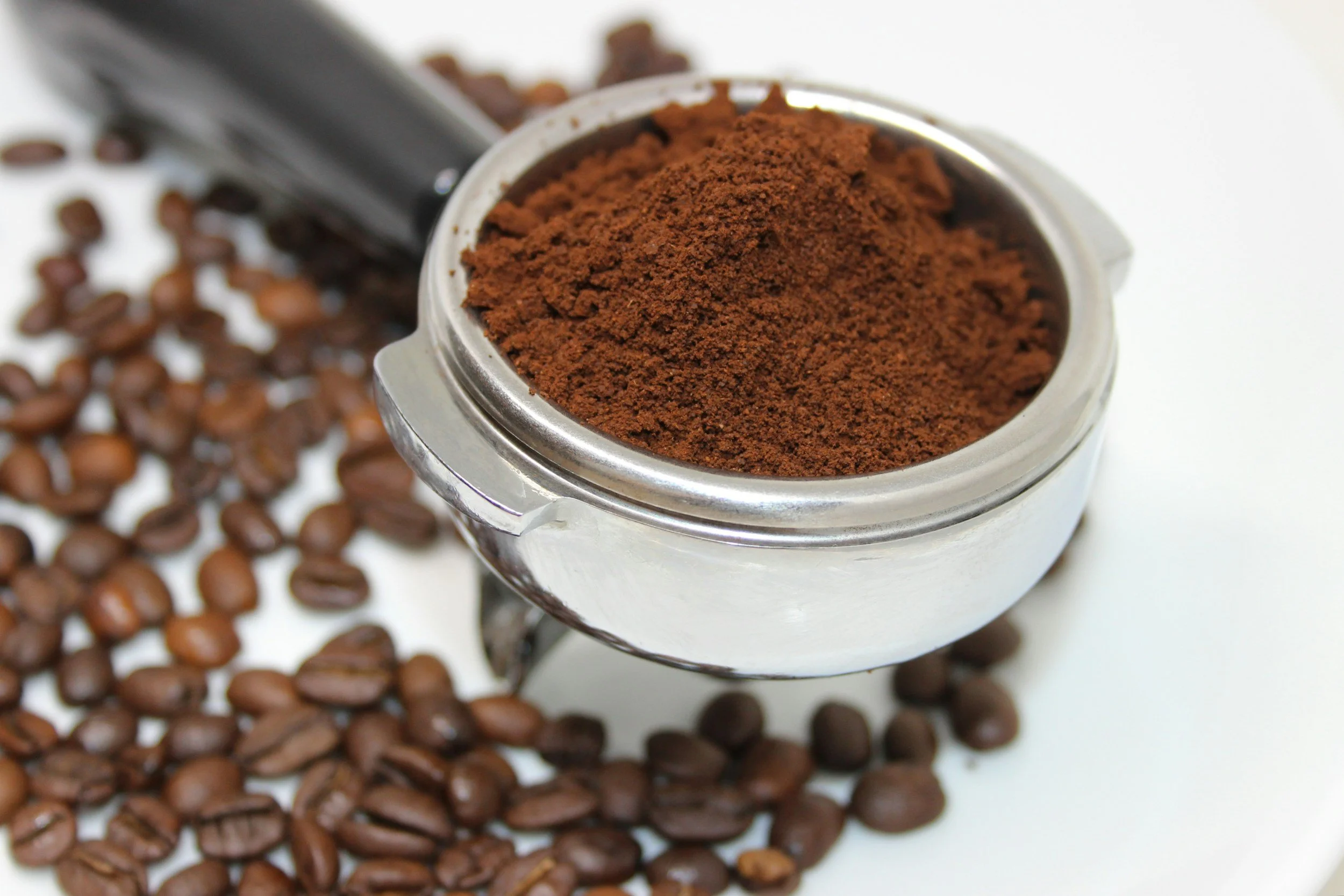 A metal container filled with ground coffee, surrounded by scattered coffee beans on a white surface.
