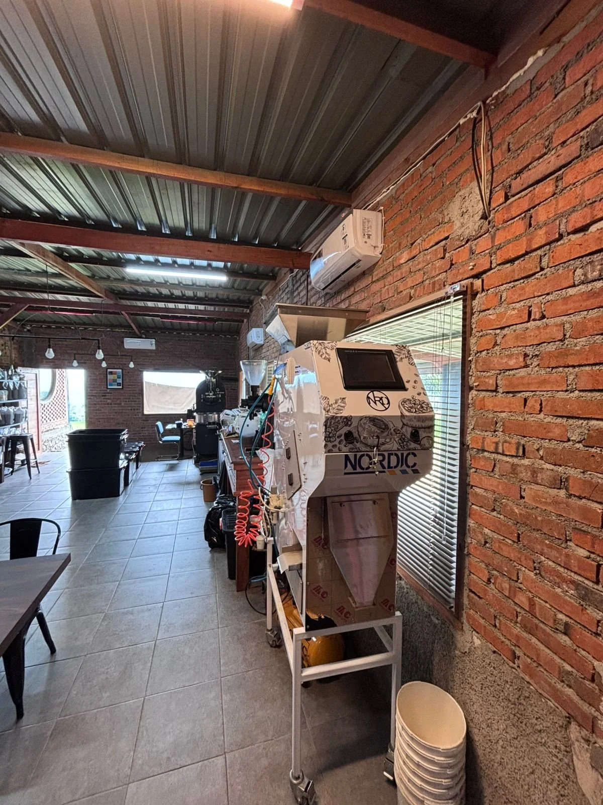 Interior of a rustic coffee shop or café with exposed red brick walls, metal ceiling, and tiled floor. There is a Nordic brand coffee bean roaster machine in the foreground, and various brewing equipment, tables, and chairs in the background.