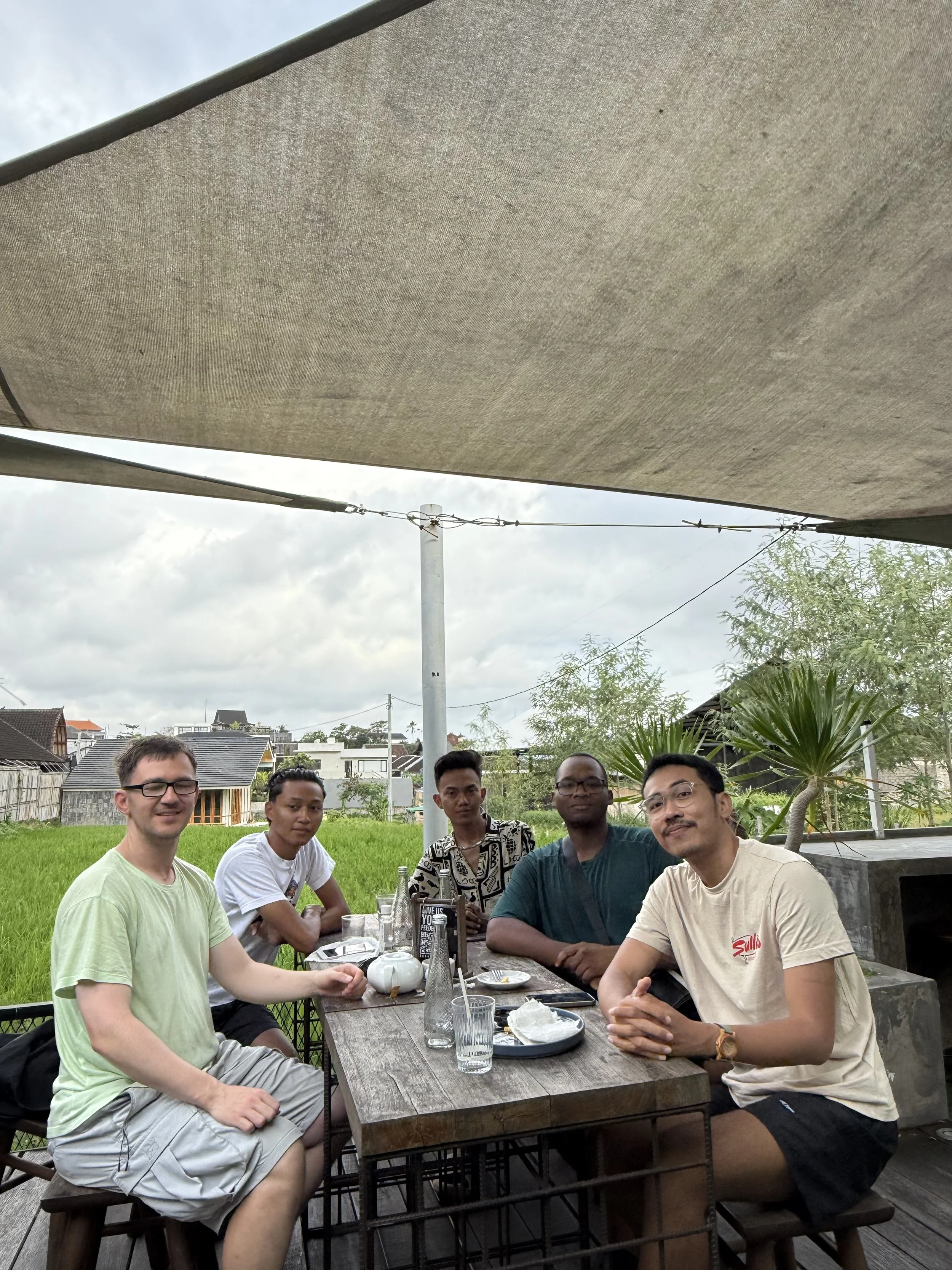 Six men sitting at an outdoor wooden dining table on a patio, with a green lawn, trees, and houses in the background, under a large beige sunshade, during daytime.