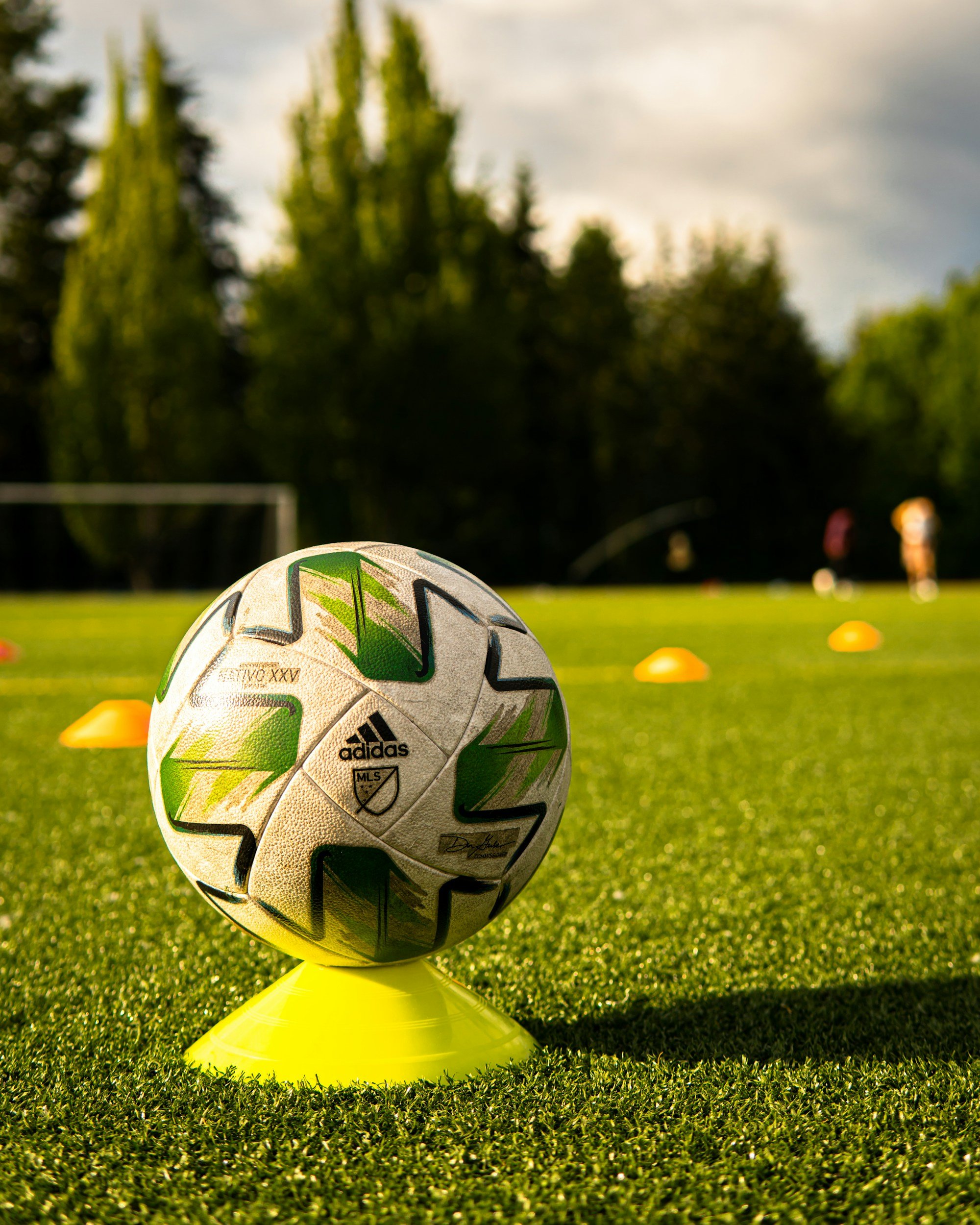 Soccer ball on a yellow cone, on a grassy field with orange cones and trees in the background.