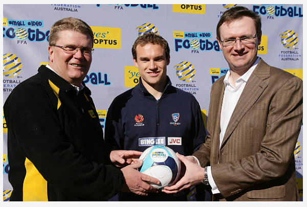 Three men standing in front of a Football Federation of Australia backdrop, holding a soccer ball together. The man on the left is wearing glasses and a black jacket with yellow accents. The man in the middle is wearing a dark sports jacket with vari