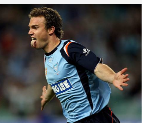 A soccer player celebrating on the field, wearing a light blue and navy uniform with a large badge on the chest.