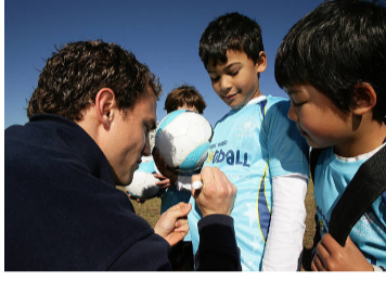 A man signing autographs for young boys wearing sports jerseys outdoors on a sunny day.