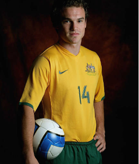 A young male soccer player wearing a yellow jersey with the number 14, holding a blue and white soccer ball, against a dark background.