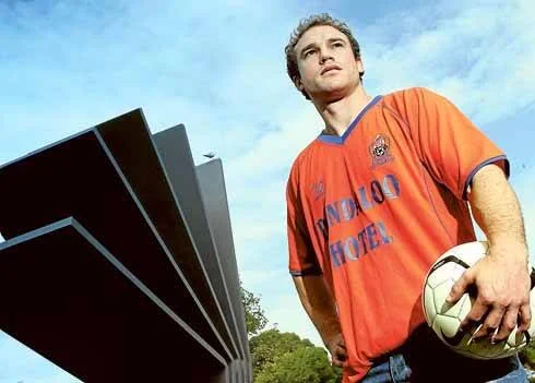 Young man in an orange sports jersey holding a soccer ball, overlooking a large trophy, outdoors on a cloudy day.