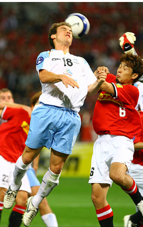 Two soccer players, one in a blue and white uniform and the other in a red and white uniform, contesting for the ball in mid-air during a match.