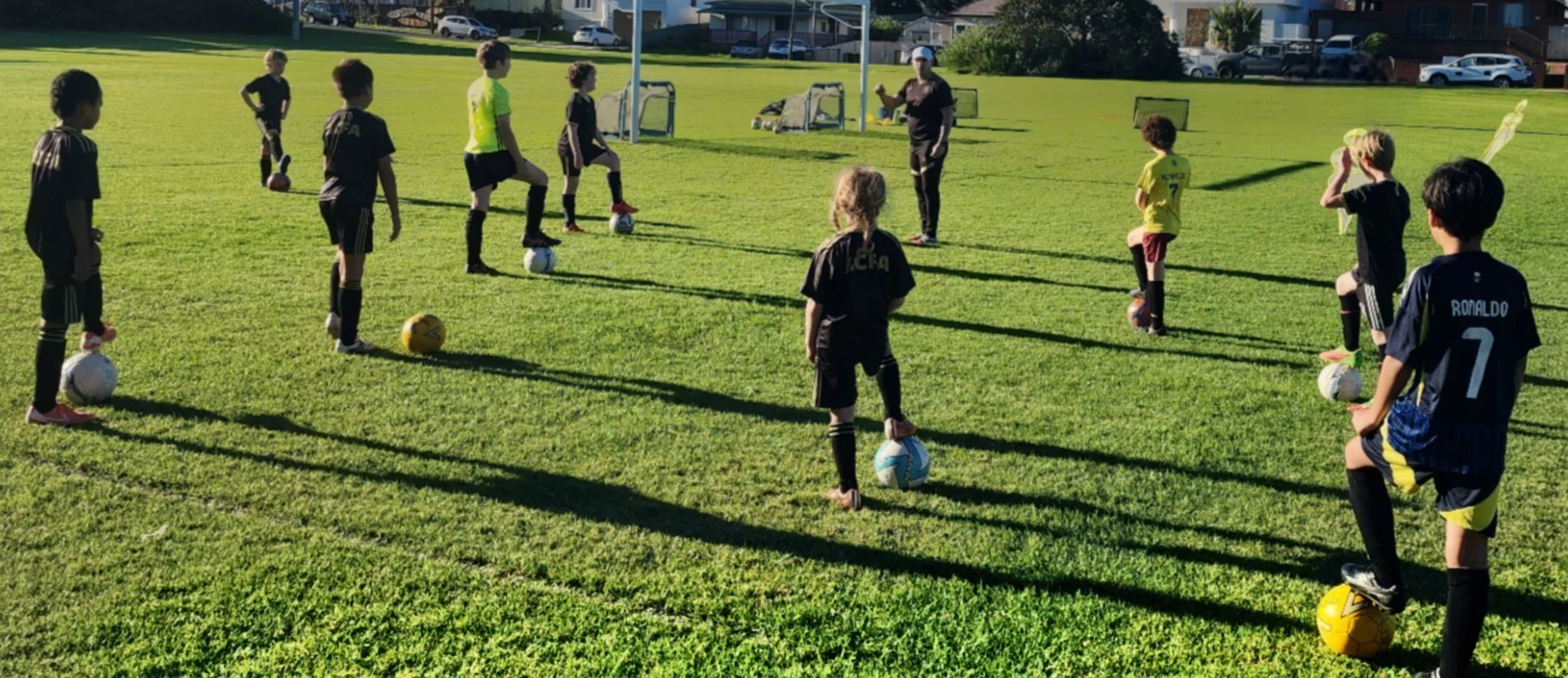 Children participating in a soccer training session on a grassy field, lined up with soccer balls at their feet, facing a coach who is giving instructions. The children wear sports uniforms, and the scene is outdoors with houses and parked cars in th