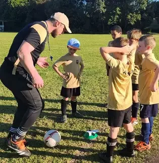 A man coaching a youth soccer team on a grassy field with children standing in a line, wearing yellow and black uniforms, and a ball on the ground.
