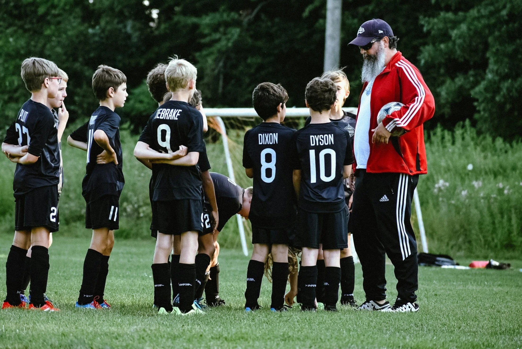 Young soccer players standing in a line listening to their coach, who is holding a soccer ball, on a green field with a goalpost and trees in the background.