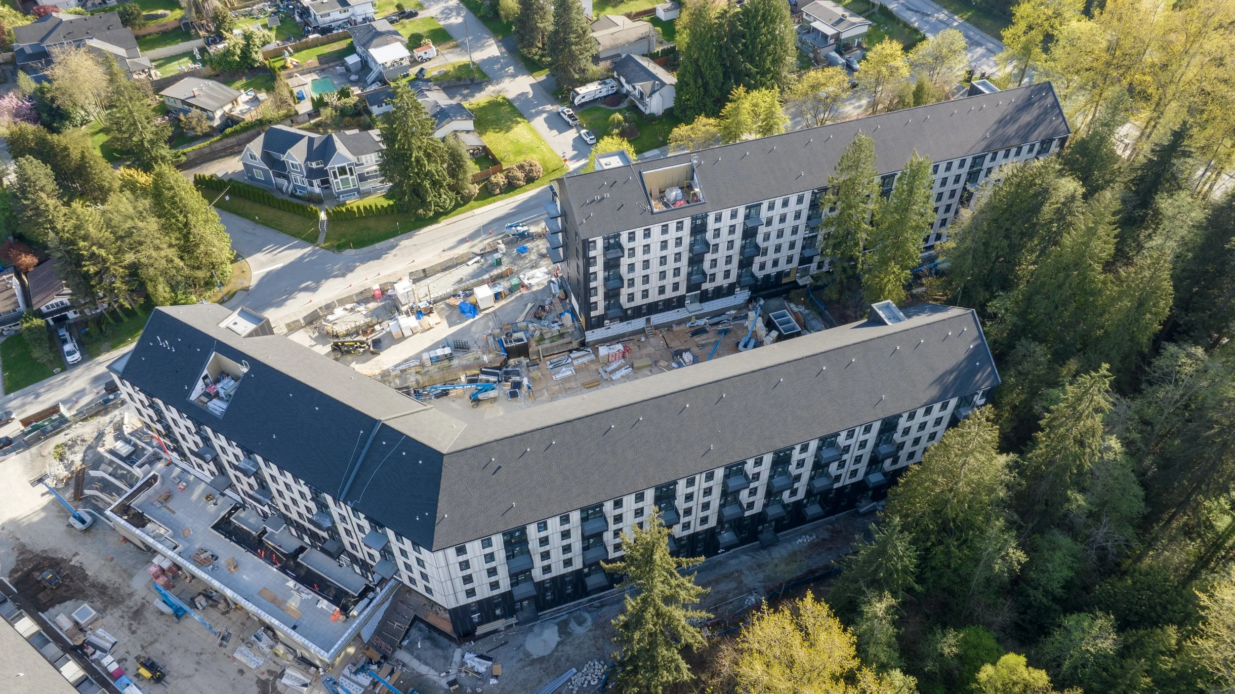 An aerial view of a large residential building under construction, with construction equipment and materials visible on the site, surrounded by trees and neighboring houses.