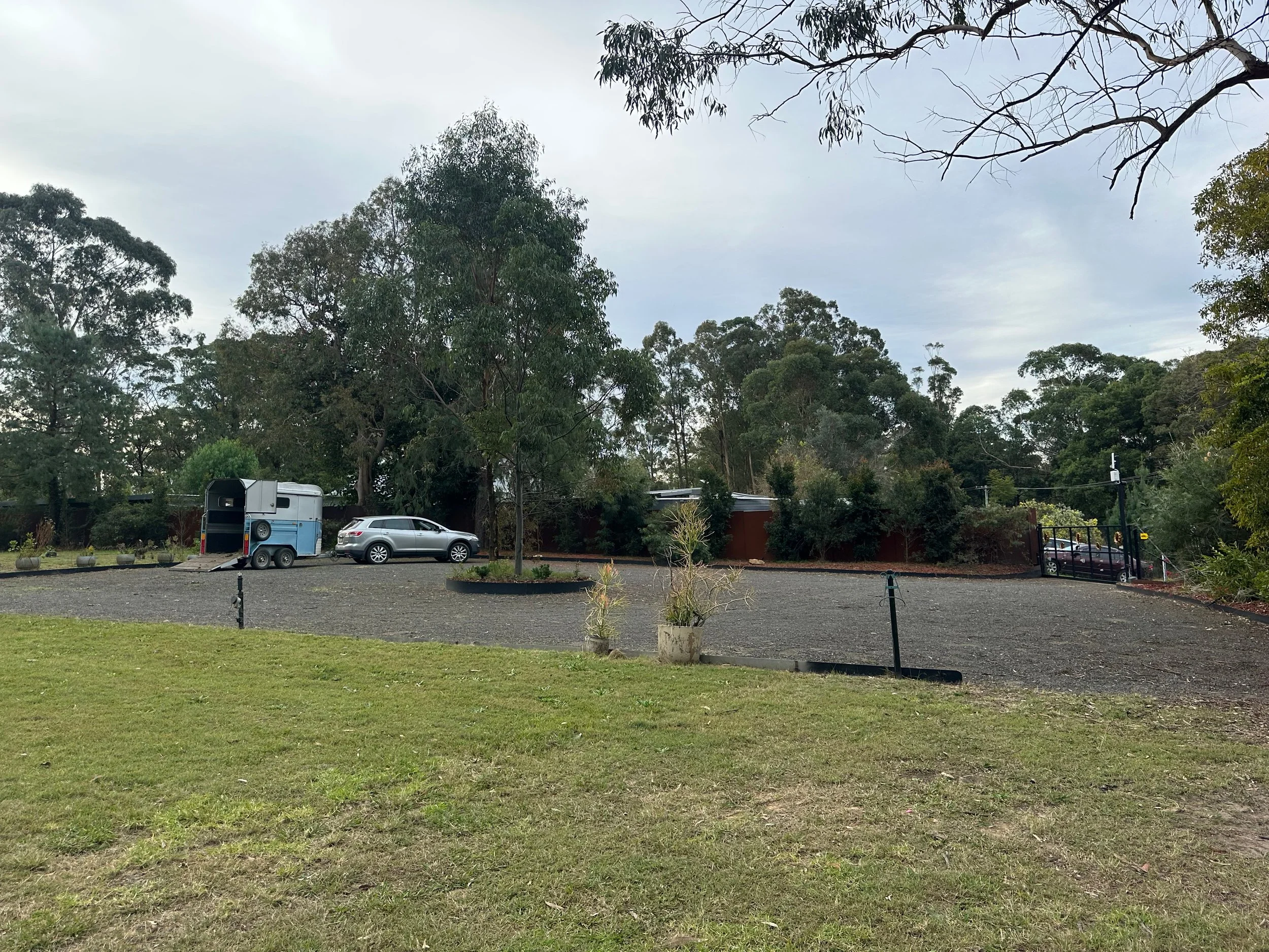 A car park surrounded by green grass, with two cars and a trailer, and a background of tall trees under a cloudy sky.