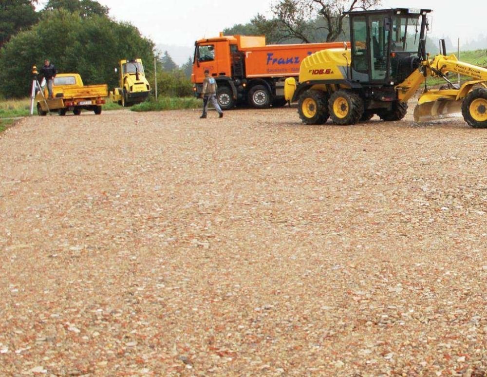 Construction site with heavy machinery including a yellow roller, an orange water truck, and a yellow grader on a gravel surface, with workers present and trees in the background.