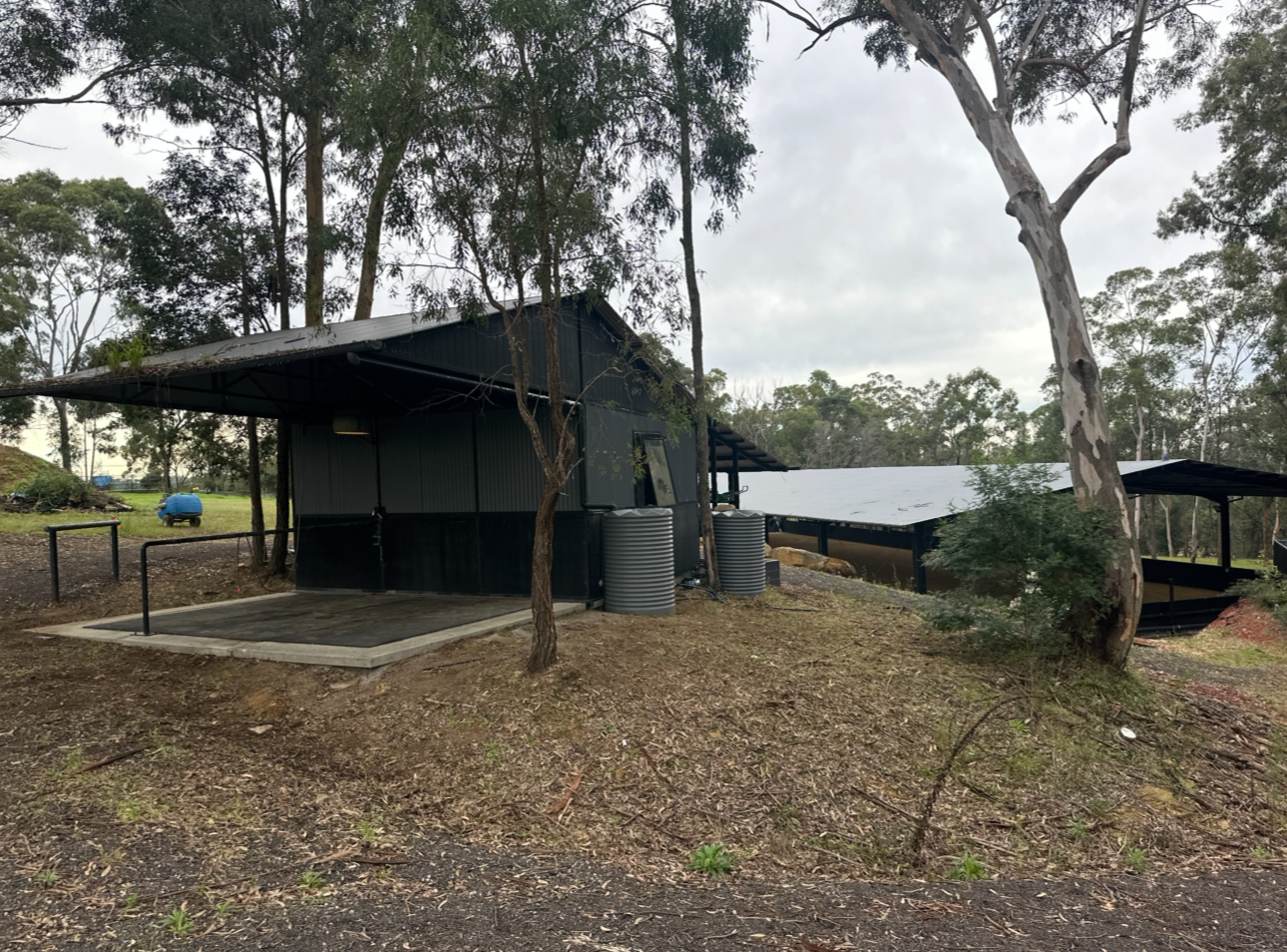 A small black metal building with a sloped roof, situated in a rural outdoor setting with trees around, featuring water tanks and a small covered area.