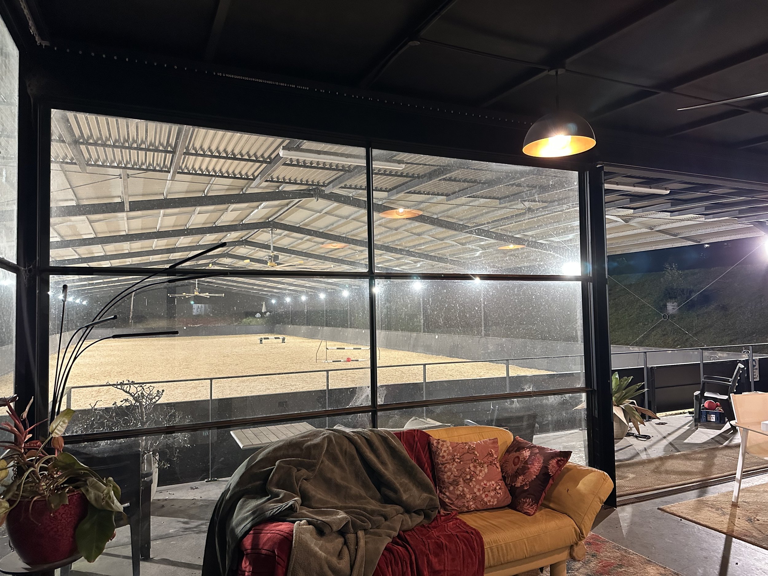 Living room with a yellow couch, cushions, jackets, and plants, seen through glass walls looking out on a sports field with a soccer goal, covered by a metal roof ceiling.