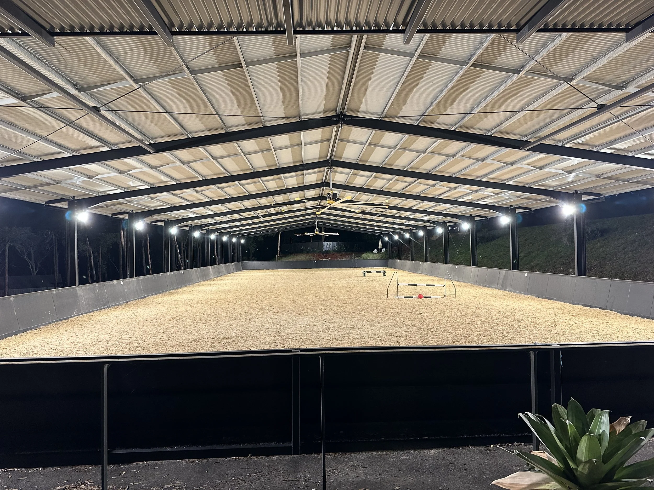 An indoor equestrian arena under a large metal roof at night with bright lights illuminating the area.