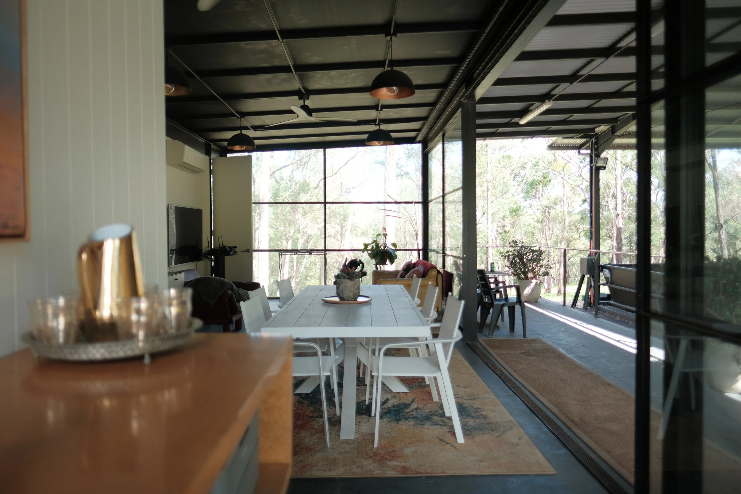 A bright indoor dining area with large windows showing trees outside. There is a white dining table with a potted plant and chairs around it, and the ceiling has hanging lights. The room has a modern, open design with a view of an outdoor area with trees.