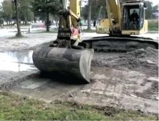 A toy excavator lifting a small pink toy truck in a park with trees and grass visible.