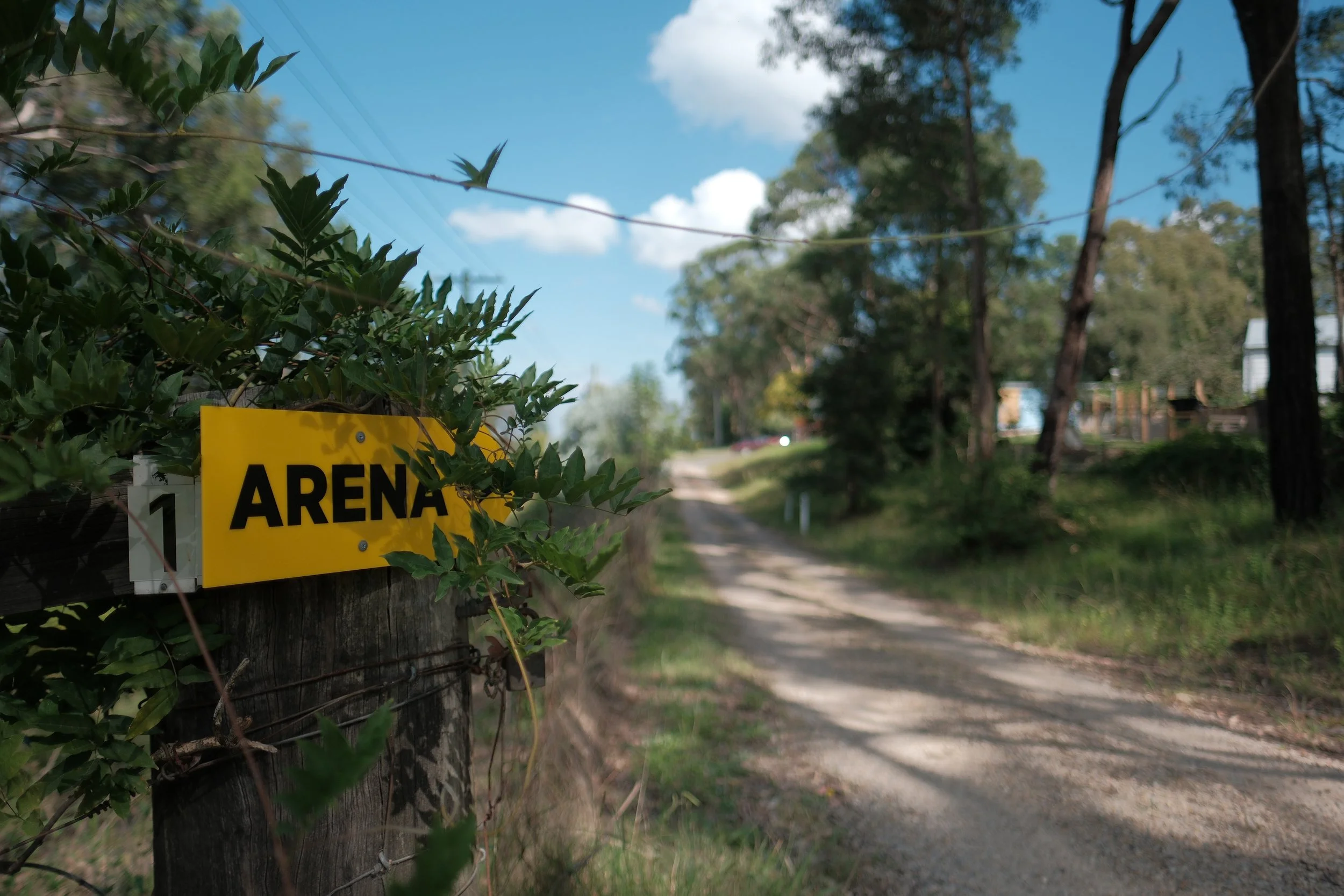 A wooden fence with a yellow sign reading 'ARENA' attached to it. Green foliage and trees are in the background along a dirt road under a partly cloudy blue sky.