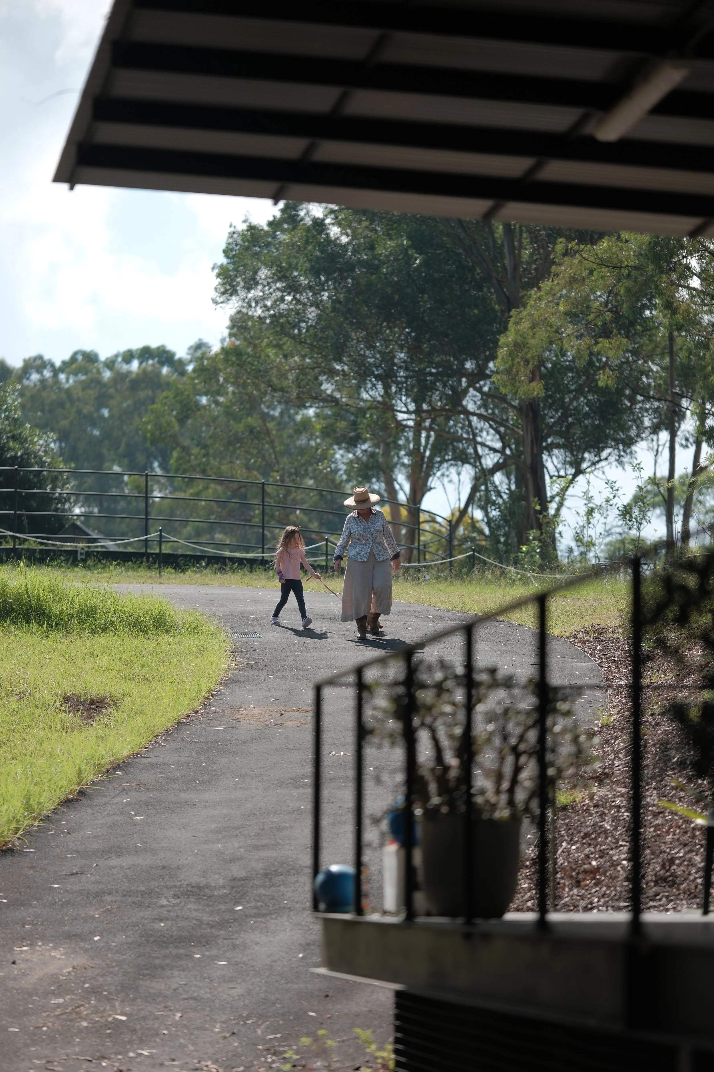 A person and a child walking on a paved path in a park, viewed from inside the indoor arena lounge. There are trees and a fence in the background, and the weather is partly cloudy.