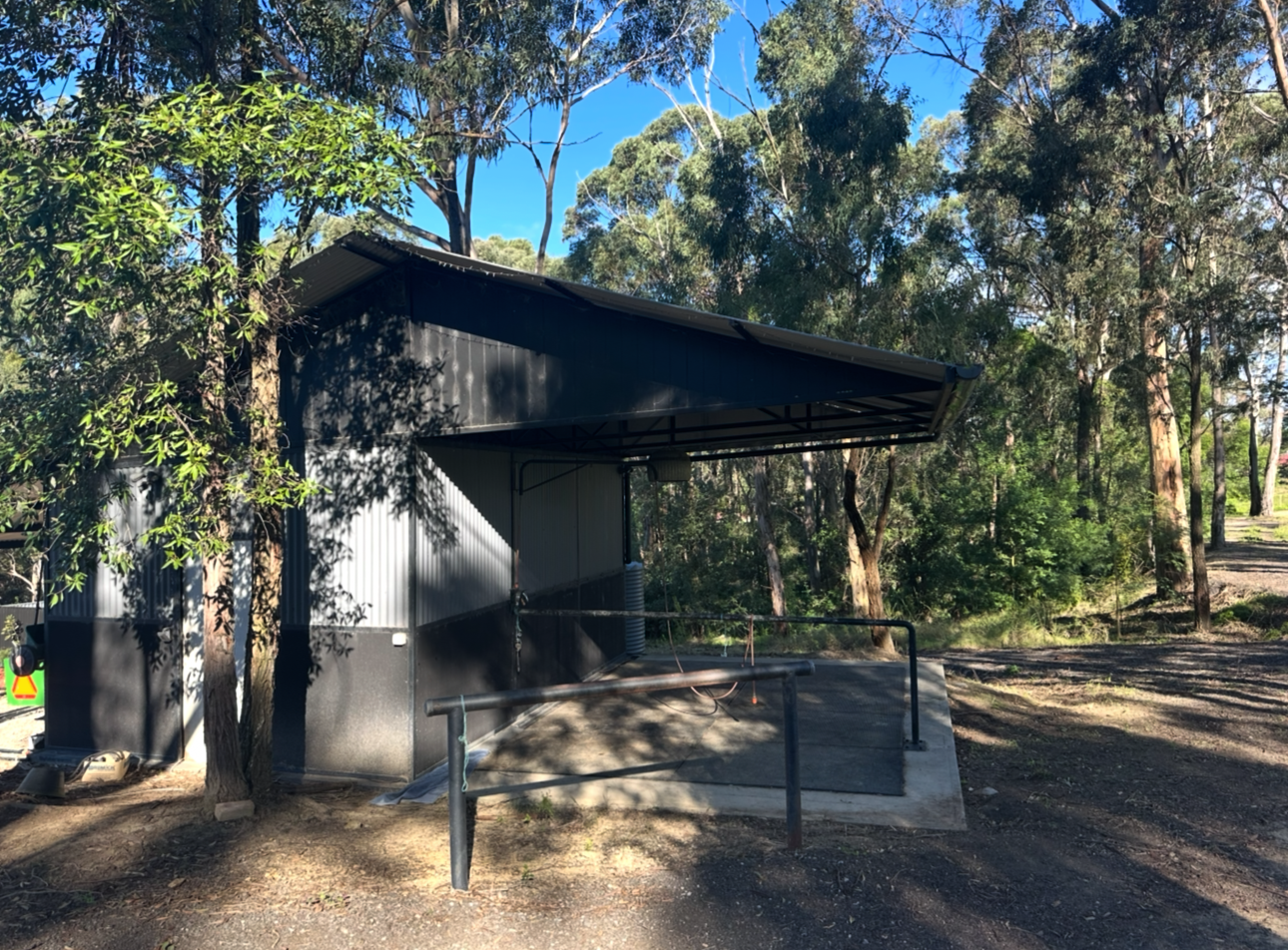 A metal wash shed with a sloped roof surrounded by trees and dirt ground in a natural setting.