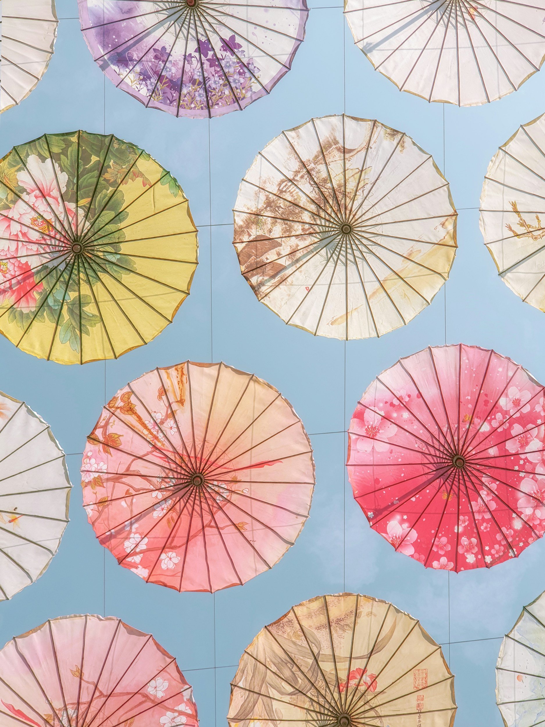 Colorful paper umbrellas with floral and nature designs are suspended in the sky against a bright blue background.