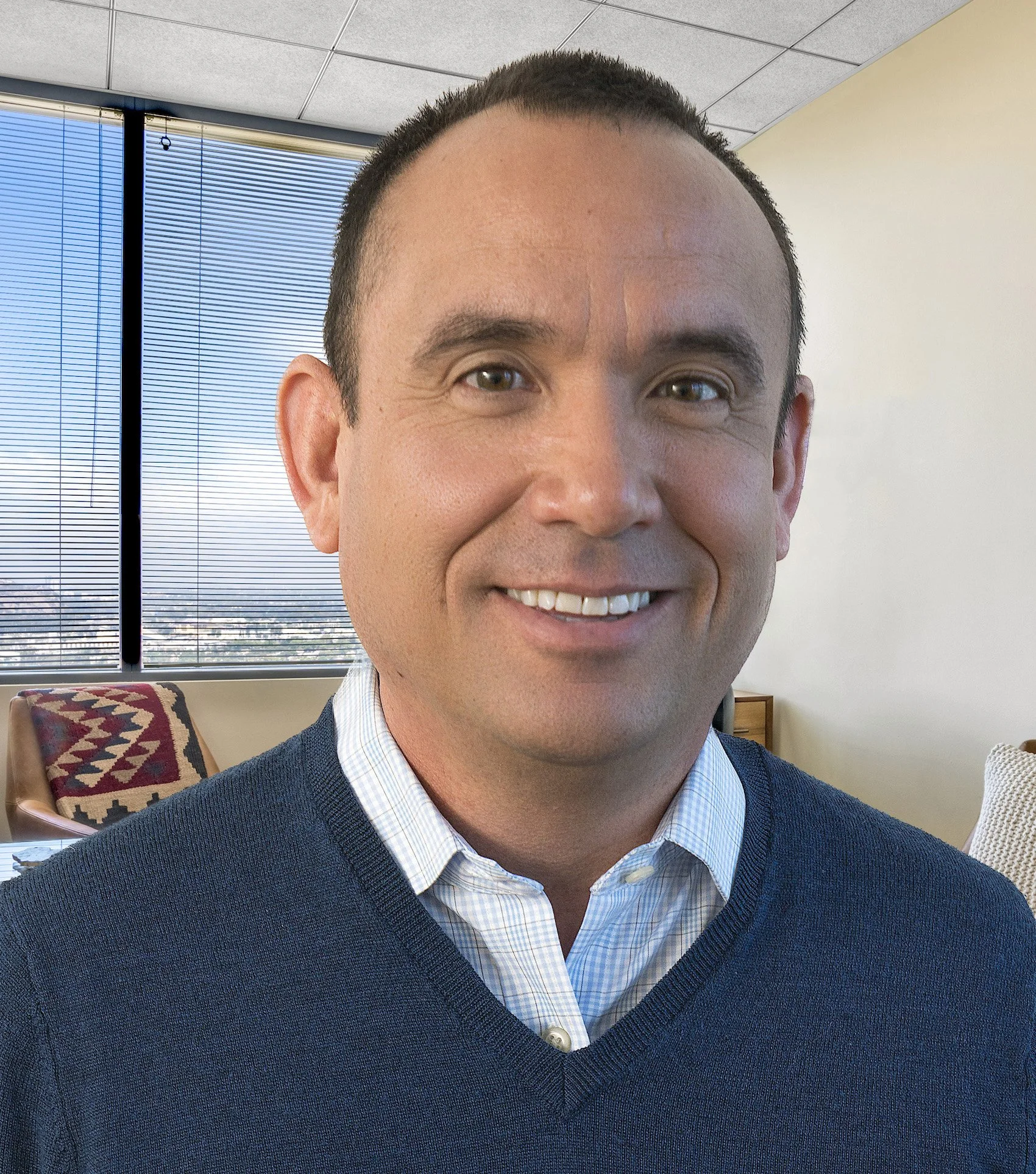 Close-up of a male therapist with short dark hair, wearing a blue shirt and a dark blazer, in an office setting with large window blinds, smiling