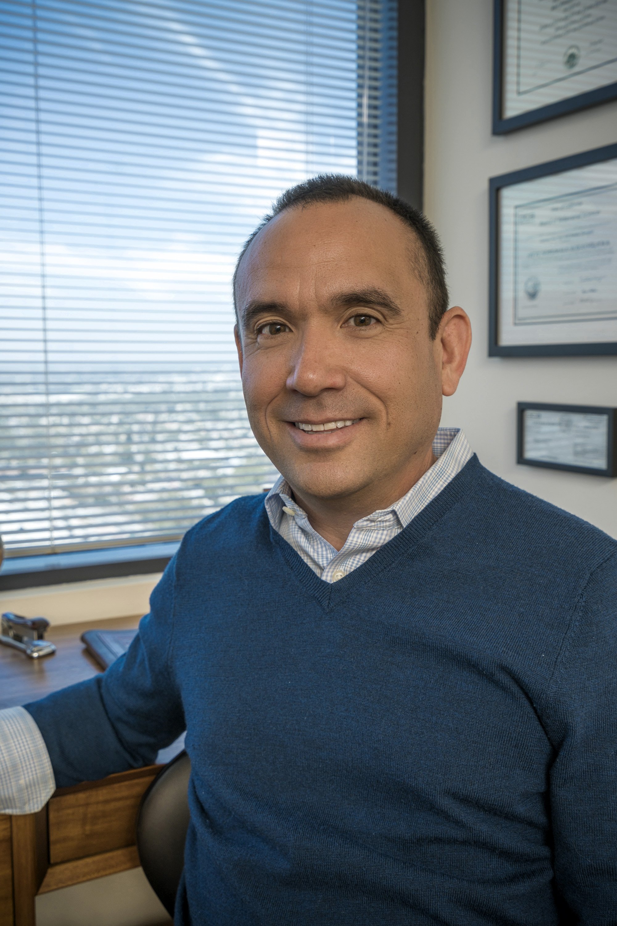 A male therapist sitting at a desk in front of a window in his office, smiling