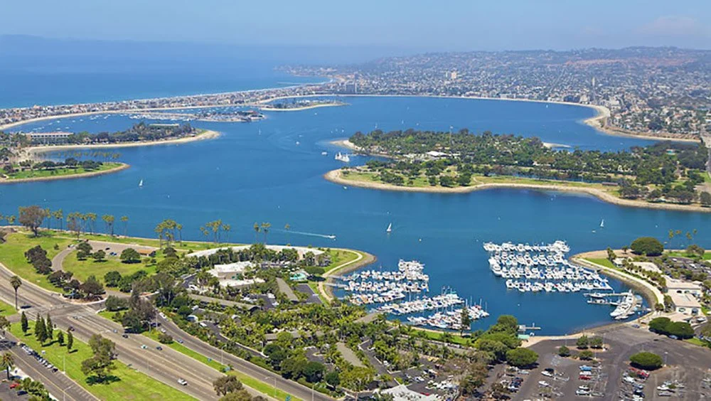 Aerial view of a harbor with boats, surrounding green parks, roads, and a cityscape in the background.