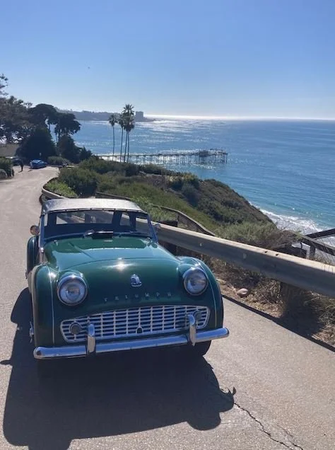 A vintage green car parked on a coastal road with the ocean and a pier in the background.