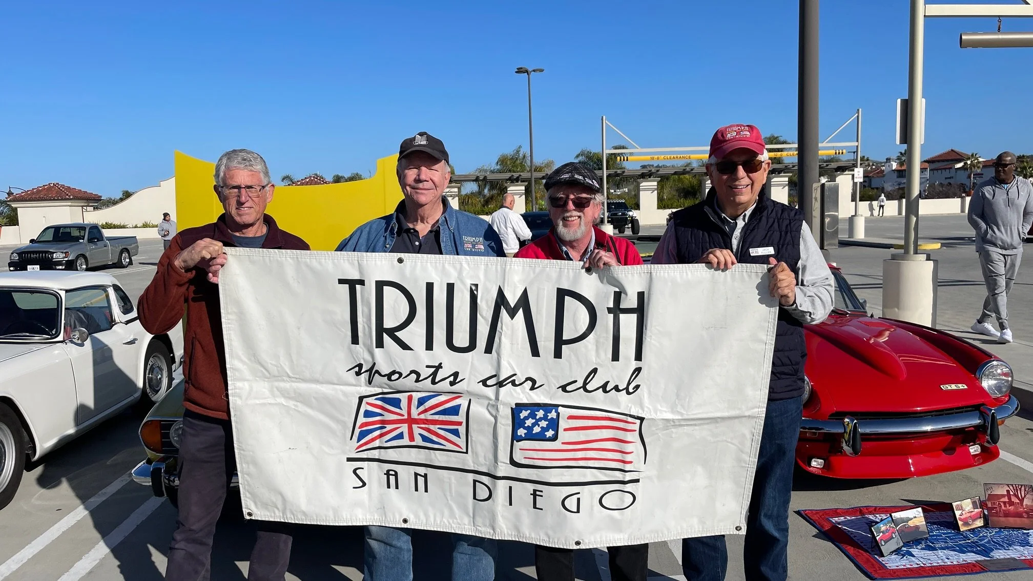 Four men holding a large white banner that reads 'Triumph sports car club' with British and American flags, standing in front of classic cars and a yellow decorative wall on a sunny day.