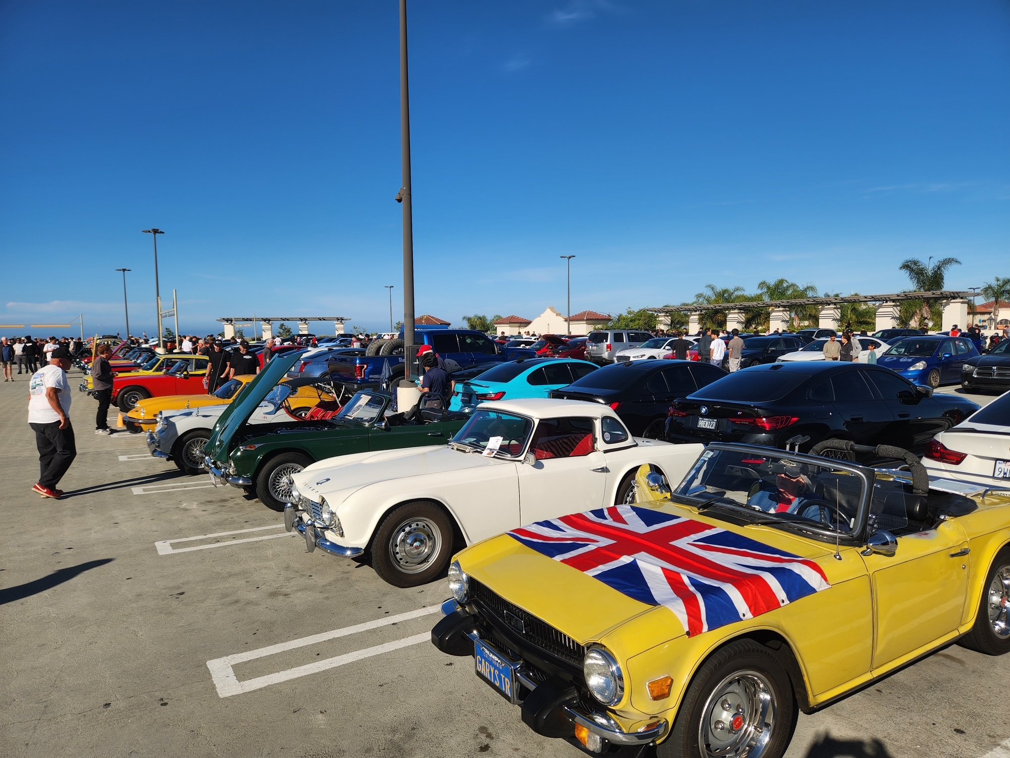 A car show in a large parking lot with a variety of vintage and classic cars, some with British flags, and many people walking around and admiring the vehicles under a clear blue sky.