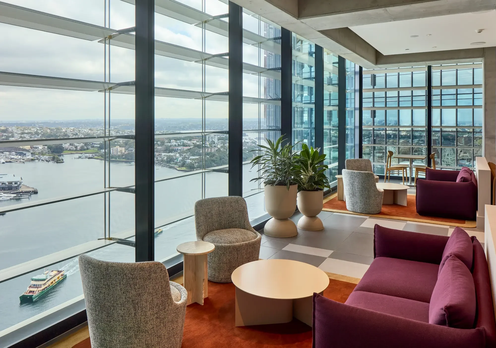 Interior view of a modern high-rise lounge area with large glass windows showing a cityscape and waterway. The space features contemporary furniture including purple sofas, armchairs, round tables, and potted plants.