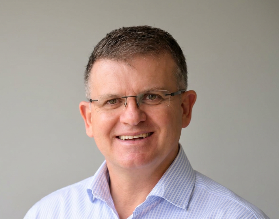 Headshot of a middle-aged man with short brown hair, glasses, and a friendly smile, wearing a light blue collared shirt against a plain light gray background.