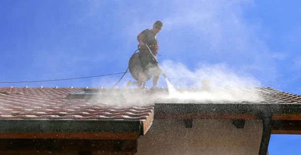 Worker power washing the roof of a house under a clear blue sky.