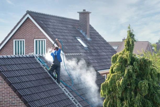 Worker pressure washing a house roof with water spray.