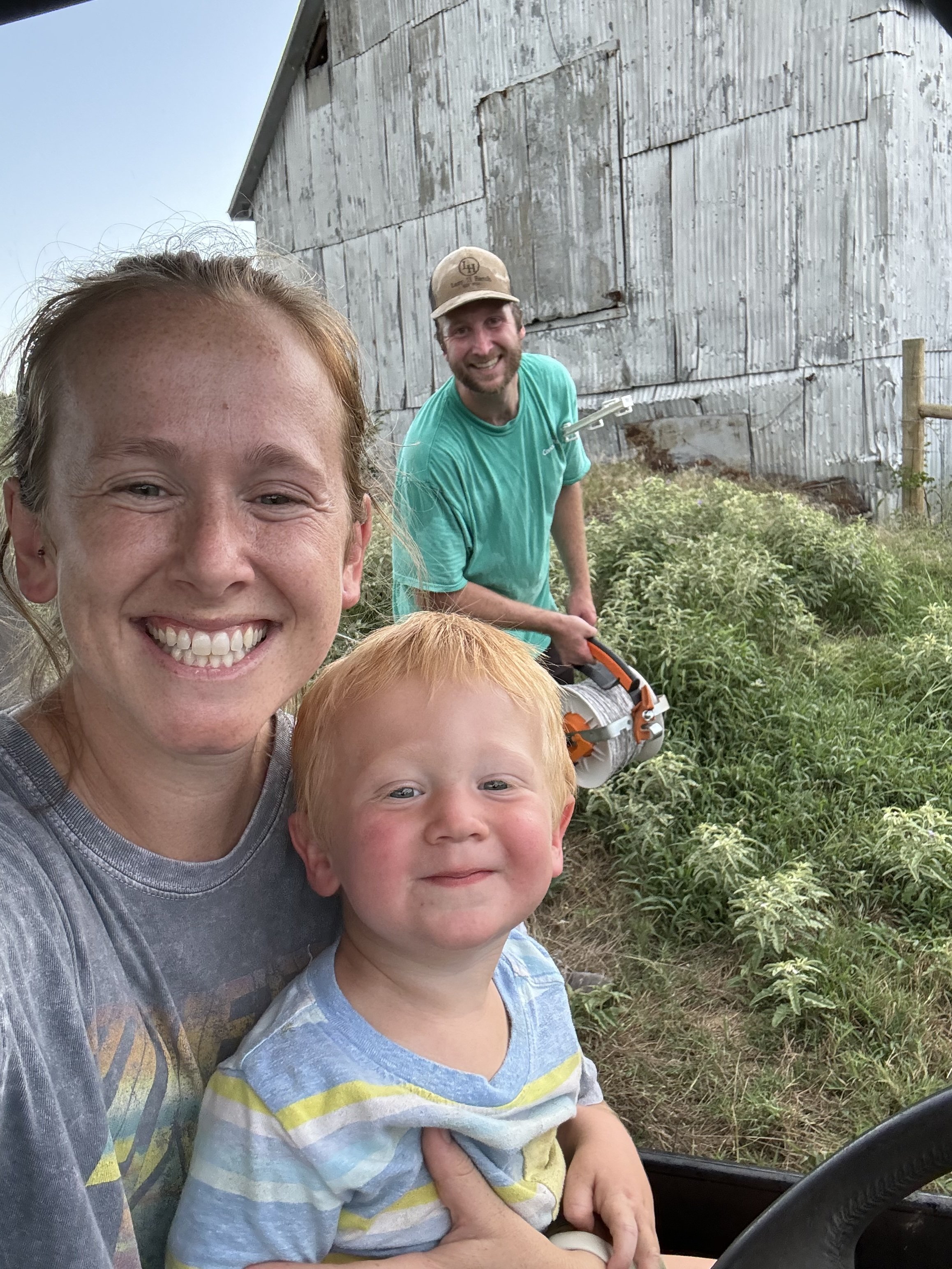 Smiling woman taking a selfie with a young boy, and a man in the background using a chainsaw, outside near a weathered barn with green plants nearby.