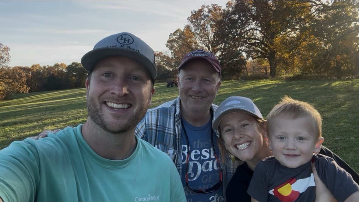 A group of four smiling people outdoors in a grassy park during sunset, including two adults, one woman, and a young boy, with trees in the background.