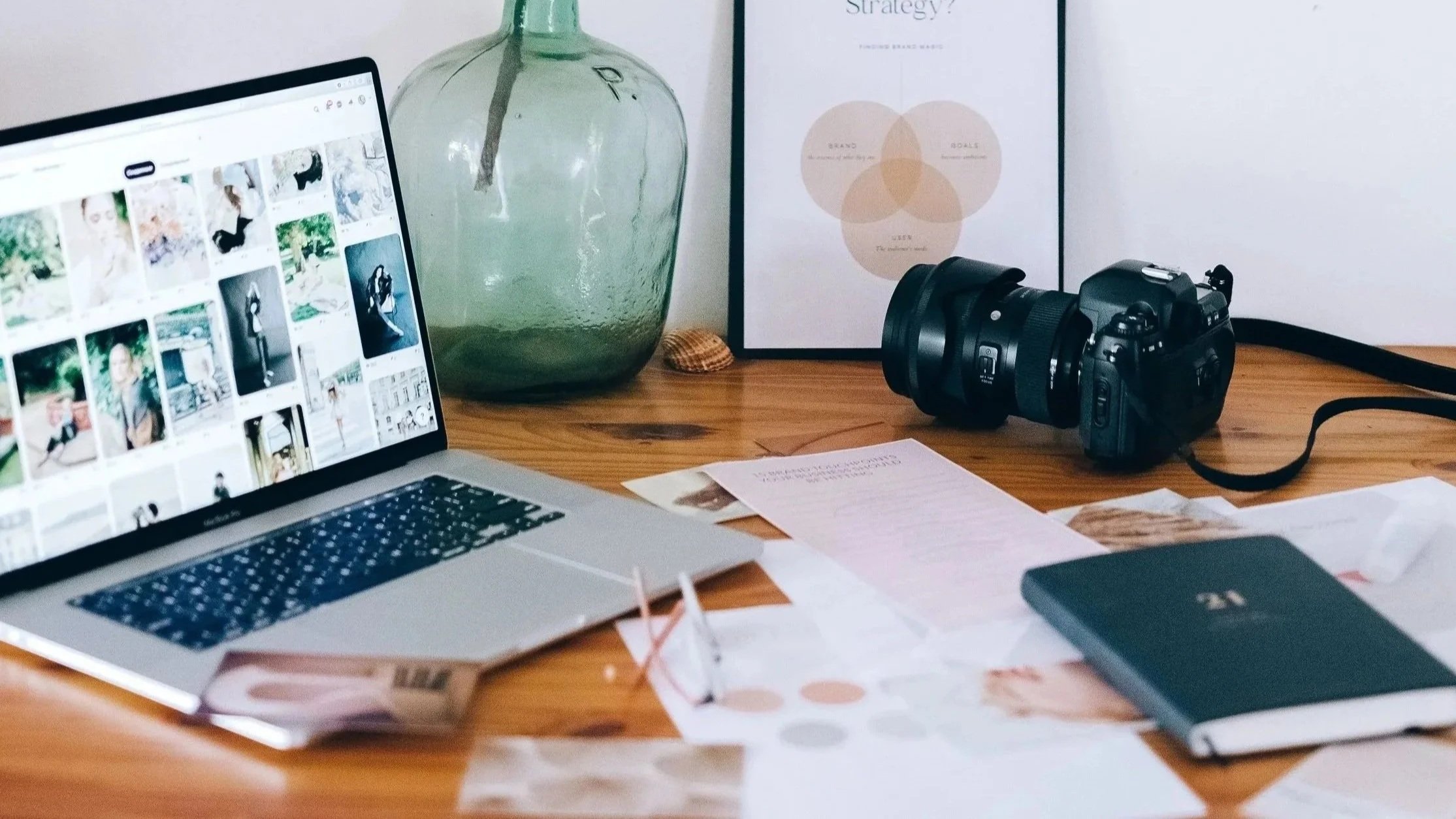 A wooden desk with a laptop displaying a photo grid, a black DSLR camera, scattered papers, a closed notebook, a large green glass vase, a framed diagram, and an orange seashell.