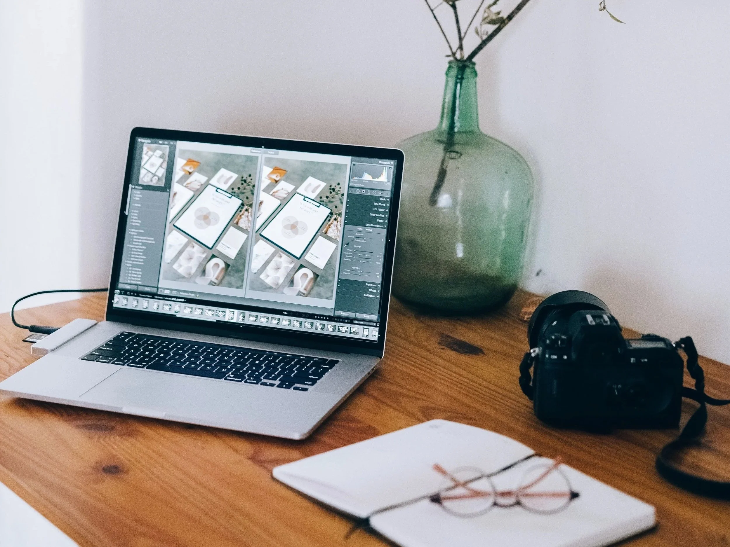 A laptop on a wooden desk displays photo editing software with multiple images of a tablet and plant on screen. To the right, a large green glass vase with branches sits against a plain wall. A camera and notebooks are also on the desk.