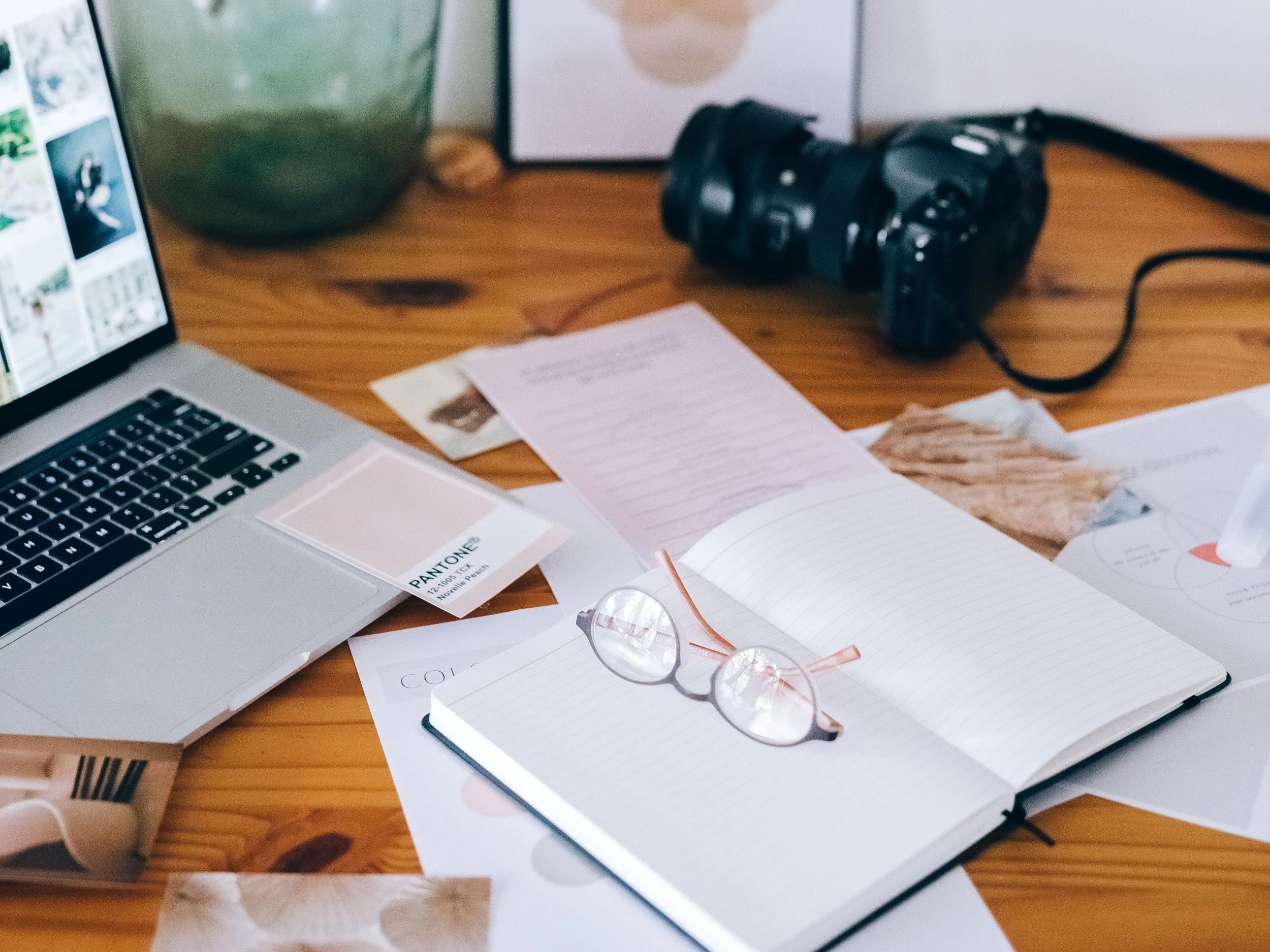 A wooden desk with a laptop, open notebooks, eyeglasses, notes, a camera, and various papers scattered on it.