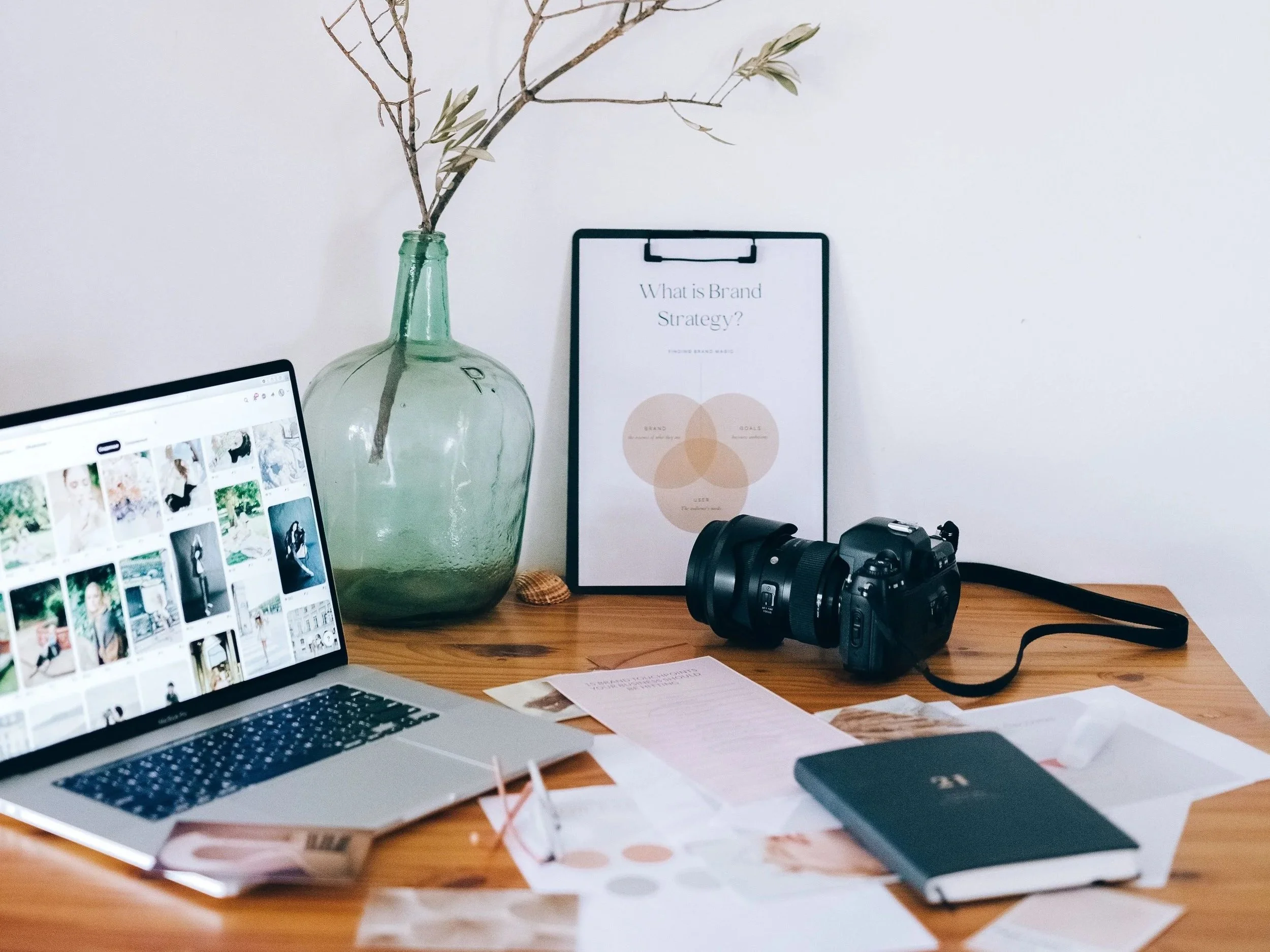A cluttered desk with a silver laptop displaying a grid of photos, a large green glass vase with bare twigs, a framed print that reads "What is Brand Strategy?", a camera on a strap, a notebook, and scattered papers and sticky notes.