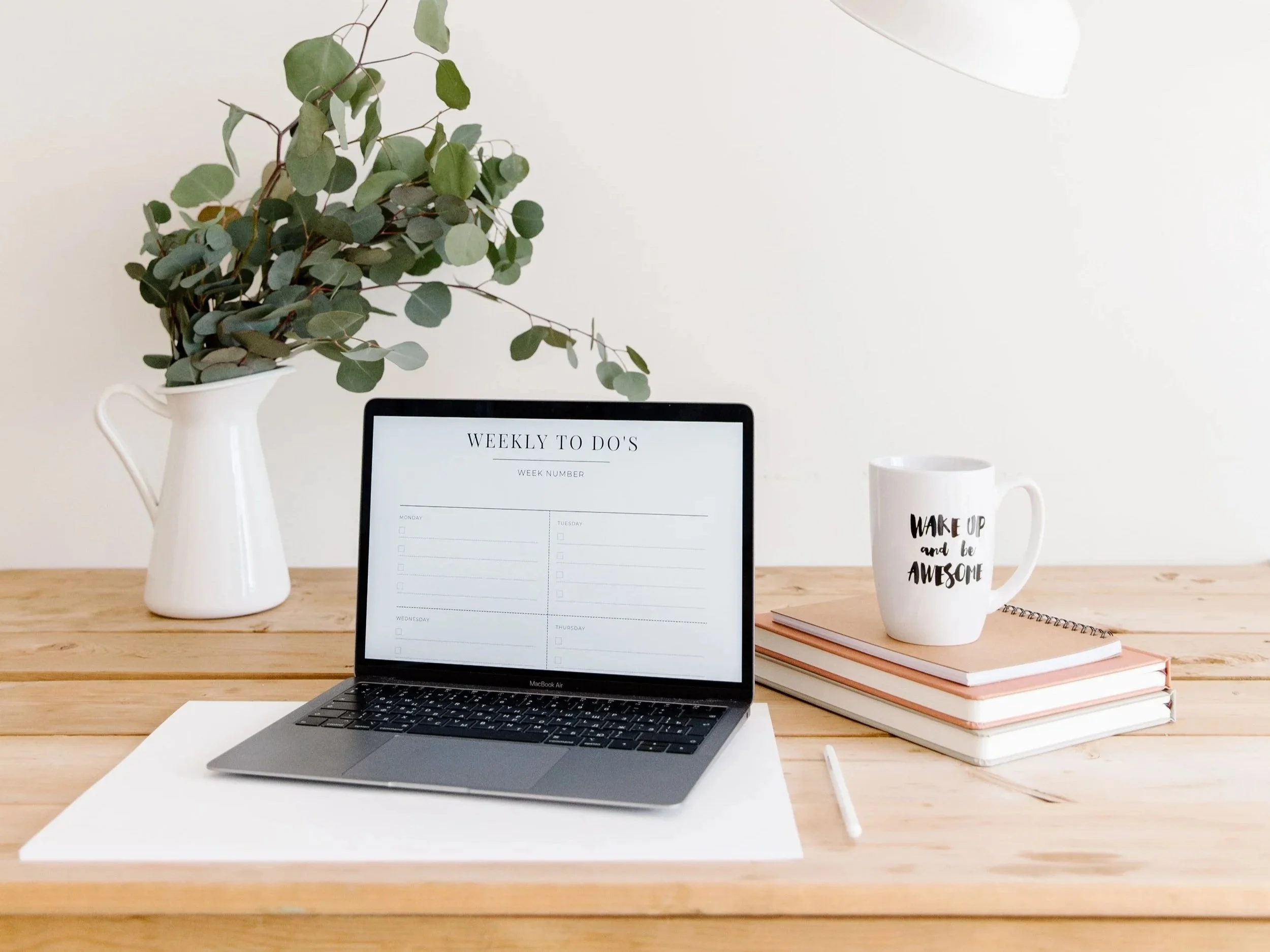 A wooden desk with a MacBook Air laptop, a white piece of paper, a white pen, a stack of pink notebooks, a white mug with black lettering that says 'Wake Up and Be Awesome', a white vase with eucalyptus branches, a digital planner titled 'Weekly To Do's', and a white lamp in the background.