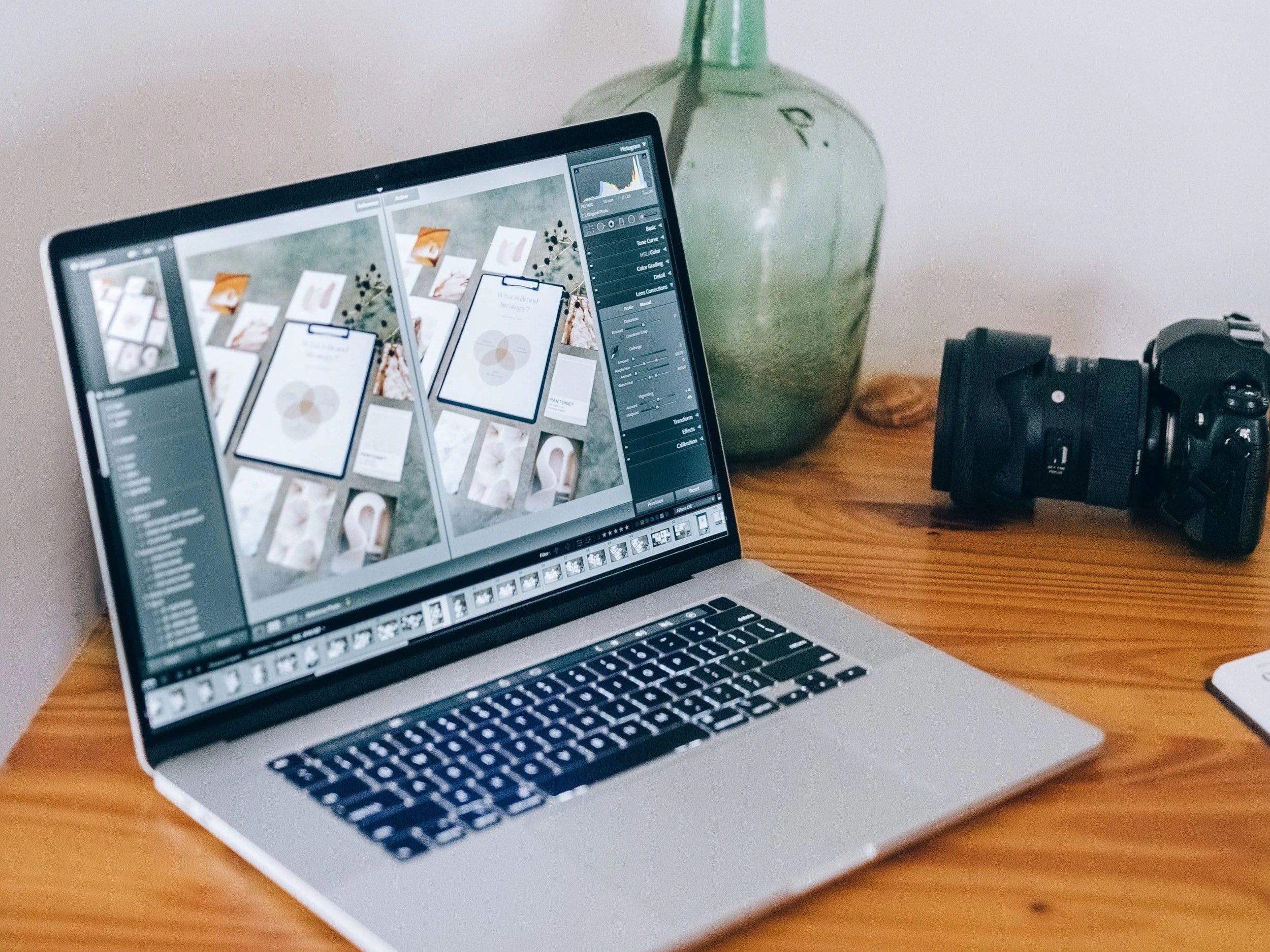 A laptop on a wooden desk displaying photo editing software, with a green glass vase and a camera nearby.