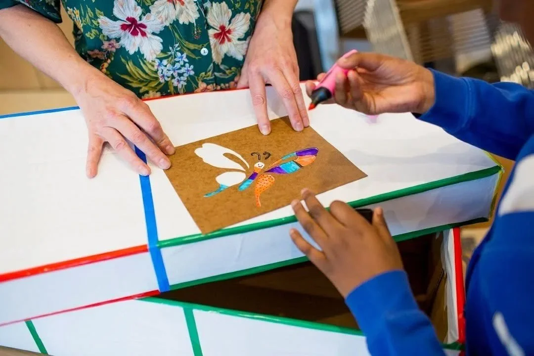 2 pairs of hands painting a cardboard eco coffin with a stencil and colourful paint