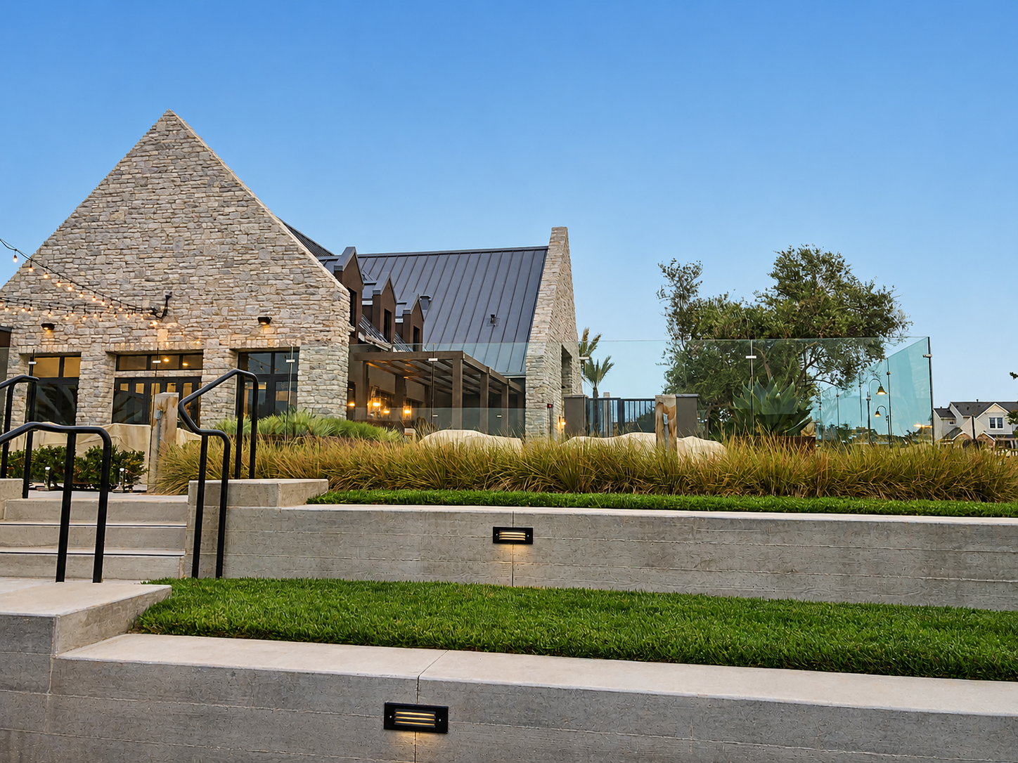 Modern building with stone and metal exterior, outdoor lighting, landscaped steps with grass and plants, large trees, and a clear sky at dusk.