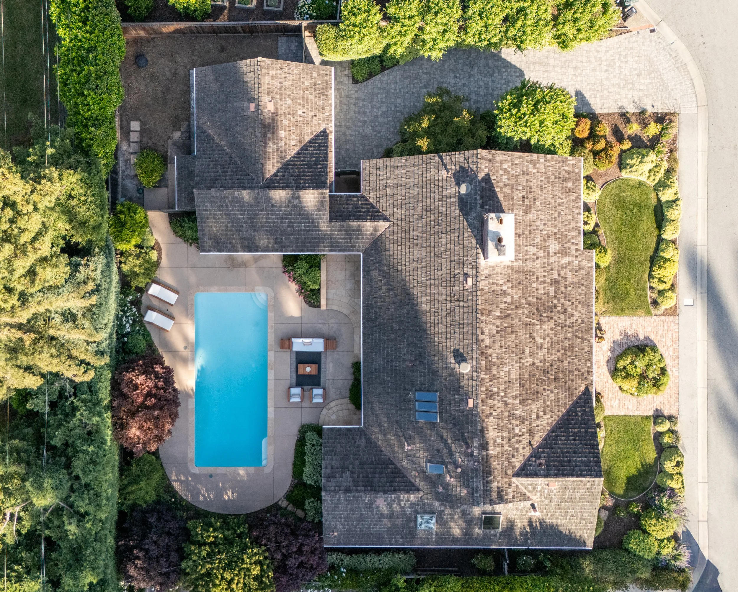 An aerial view of a suburban house with a swimming pool in the backyard, surrounded by green trees and well-maintained landscaping.