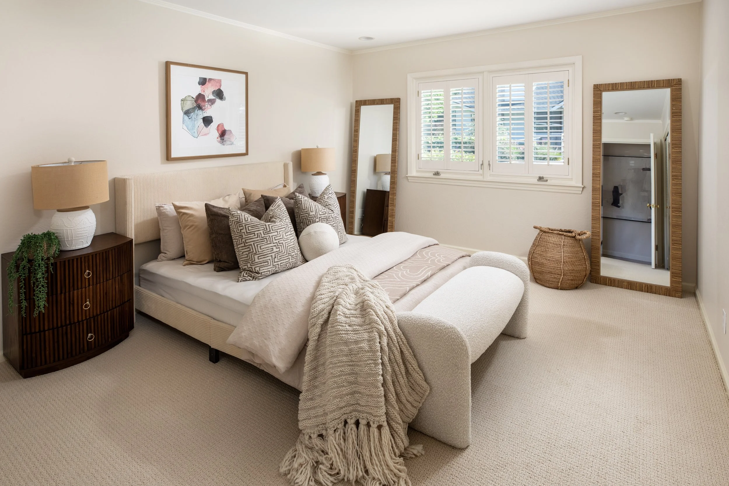 A bedroom with a cream-colored bed, multiple pillows, and a blanket. There are two bedside lamps, a mirror in a wicker frame, a framed abstract artwork, and a window with white shutters. A basket is on the floor next to the mirror, and a door leading