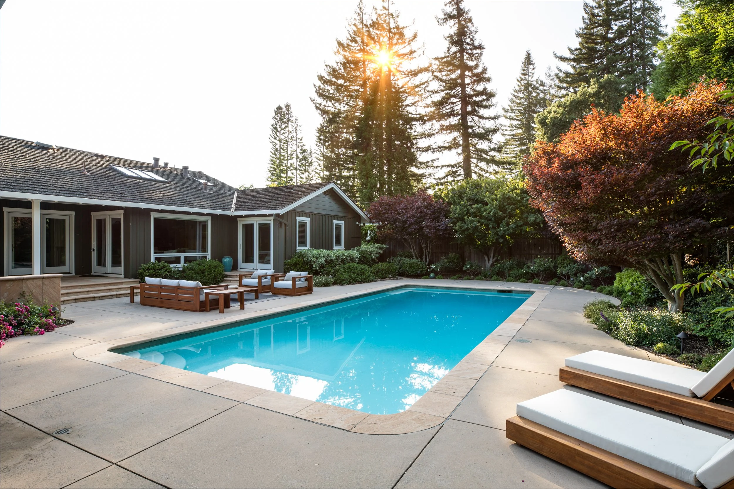A backyard with a swimming pool, surrounded by patio stones, with outdoor furniture including a long wooden table and chairs, and lounge chairs with cushions. There is a house with a gray exterior and skylights on the roof, and lush trees including t