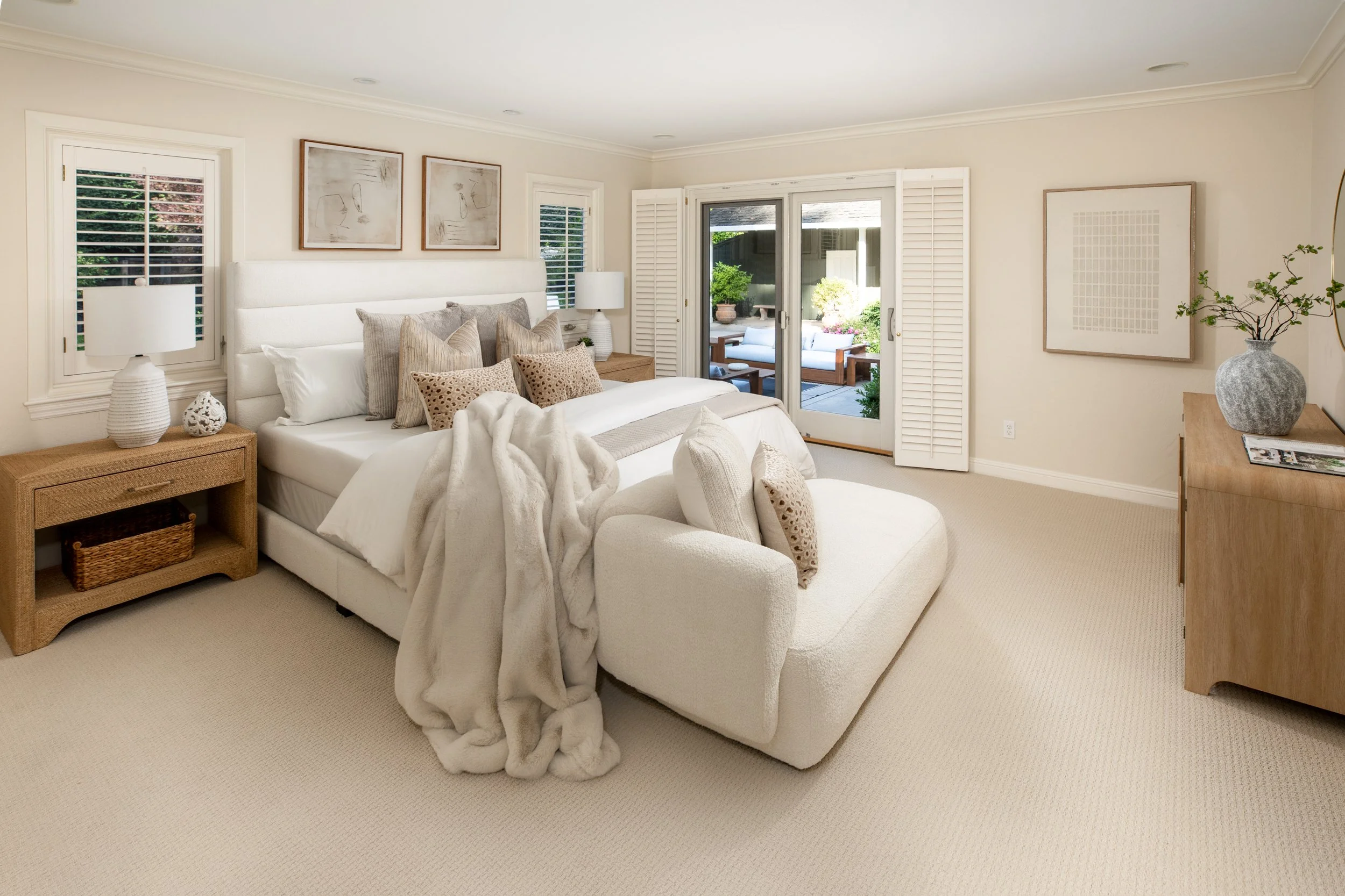 A bedroom with a white bed, beige pillows, and a cozy blanket, wooden nightstands with lamps, artwork above the bed, sliding glass door leading to an outdoor patio with seating, and a vase with branches on a wooden dresser.