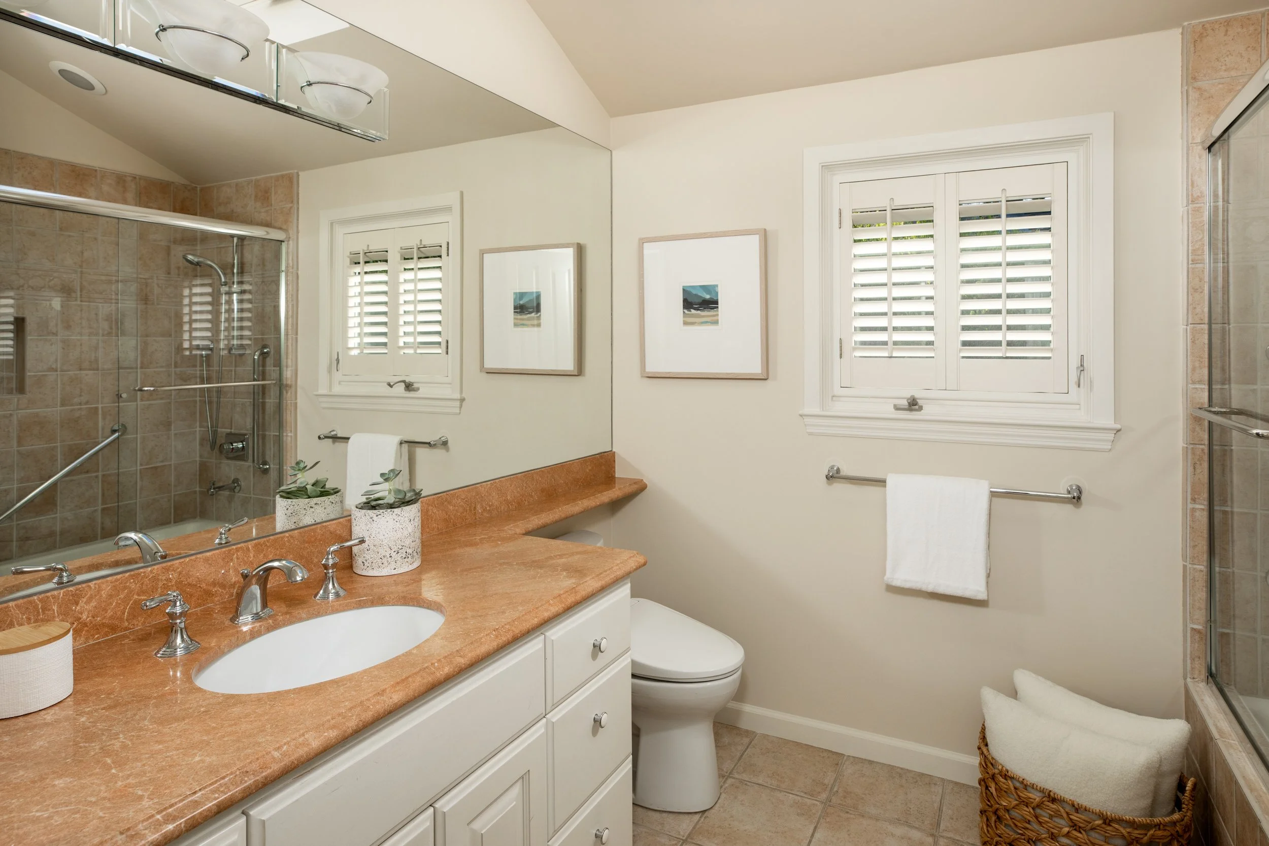 Bathroom with beige tiled shower, large mirror, white vanity with dual sinks, two small potted plants, two framed landscape paintings, four windows with white shutters, toilet, woven basket with white towels, and beige tiled floor.